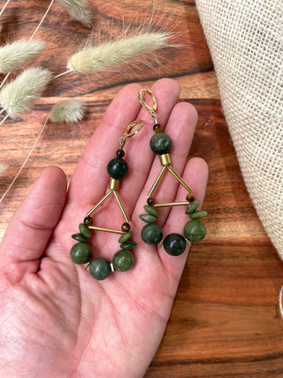 Hand holding a pair of green beaded earrings on a wooden surface with dried grasses.