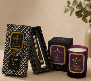 Harlem Candle Co. 'Soothing' scented candle set, featuring a purple glass jar candle, its black patterned outer box, and an open inner box displaying a gold-toned metal candle stick, arranged on a beige surface with a neutral background and a blurred plant.