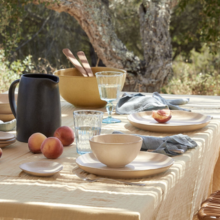 Outdoor table setting with ceramic dishes, glasses, and peaches on a striped tablecloth.