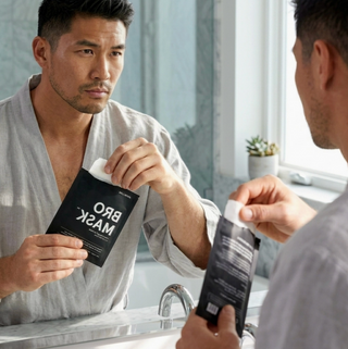 Man holding a black face mask package labeled 'BRO MASK' in front of a mirror.