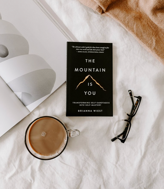 Book titled 'The Mountain is You' by Brianna Wiest with a cup of coffee and glasses on a white surface.